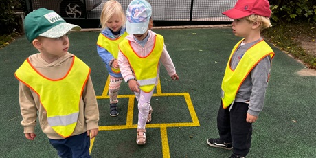 Powiększ grafikę: Czworo dzieci w kamizelkach odblaskowych gra w klasy w ogrodzie przedszkolnym